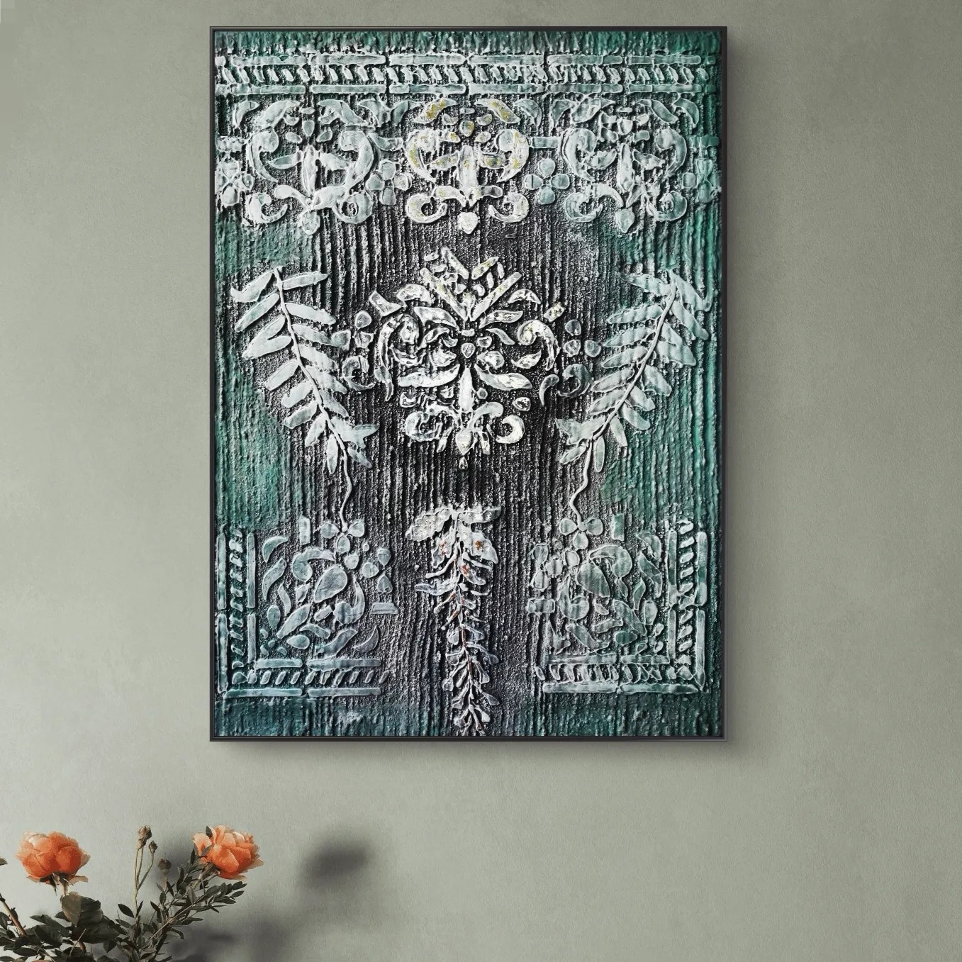 Teksturowany obraz w czarnej ramie z motywami roślinnymi i ornamentami w odcieniach zieleni i bieli, wiszący na szarej ścianie, z kwiatami w rogu.