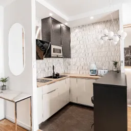 Nowoczesna kuchnia z jasnymi szafkami dolnymi i ciemnymi górnymi, drewnianym blatem, mozaiką na ścianie, czarnym zlewem i baterią, oświetlona wiszącą lampą kulistą.