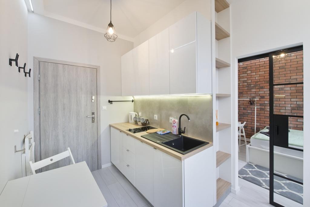 Nowoczesna, mała kuchnia w jasnych barwach z białymi, lakierowanymi szafkami, drewnianym blatem i czarnym zlewem, oświetlona listwą LED, widok na pokój z ceglaną ścianą przez przeszklone drzwi.