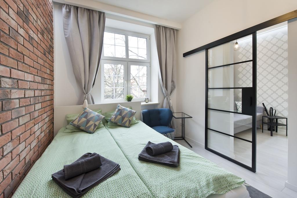 Jasna sypialnia z ceglaną ścianą, dwuosobowym łóżkiem z zieloną pościelą, przesuwnymi drzwiami w czarnej ramie i widokiem na drugi pokój.