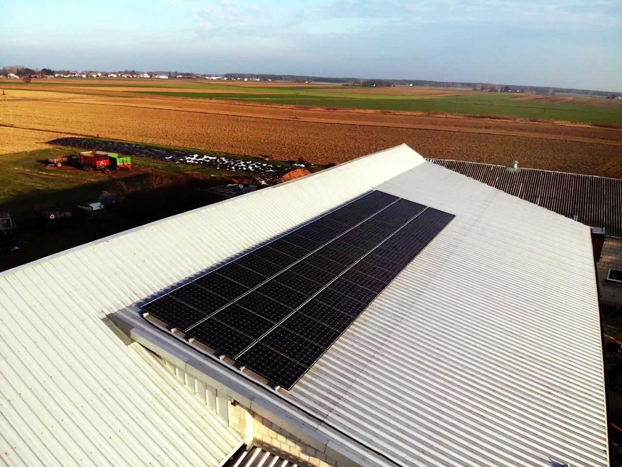 Panel słoneczny na dachu budynku gospodarczego z blachy trapezowej, widok z lotu ptaka na tle pól i zabudowań.