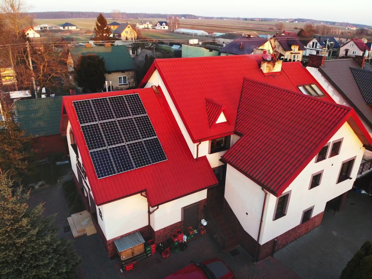 Widok z lotu ptaka na dom z czerwoną dachówką i panelami słonecznymi na dachu, otoczony zabudową jednorodzinną i polami w tle.