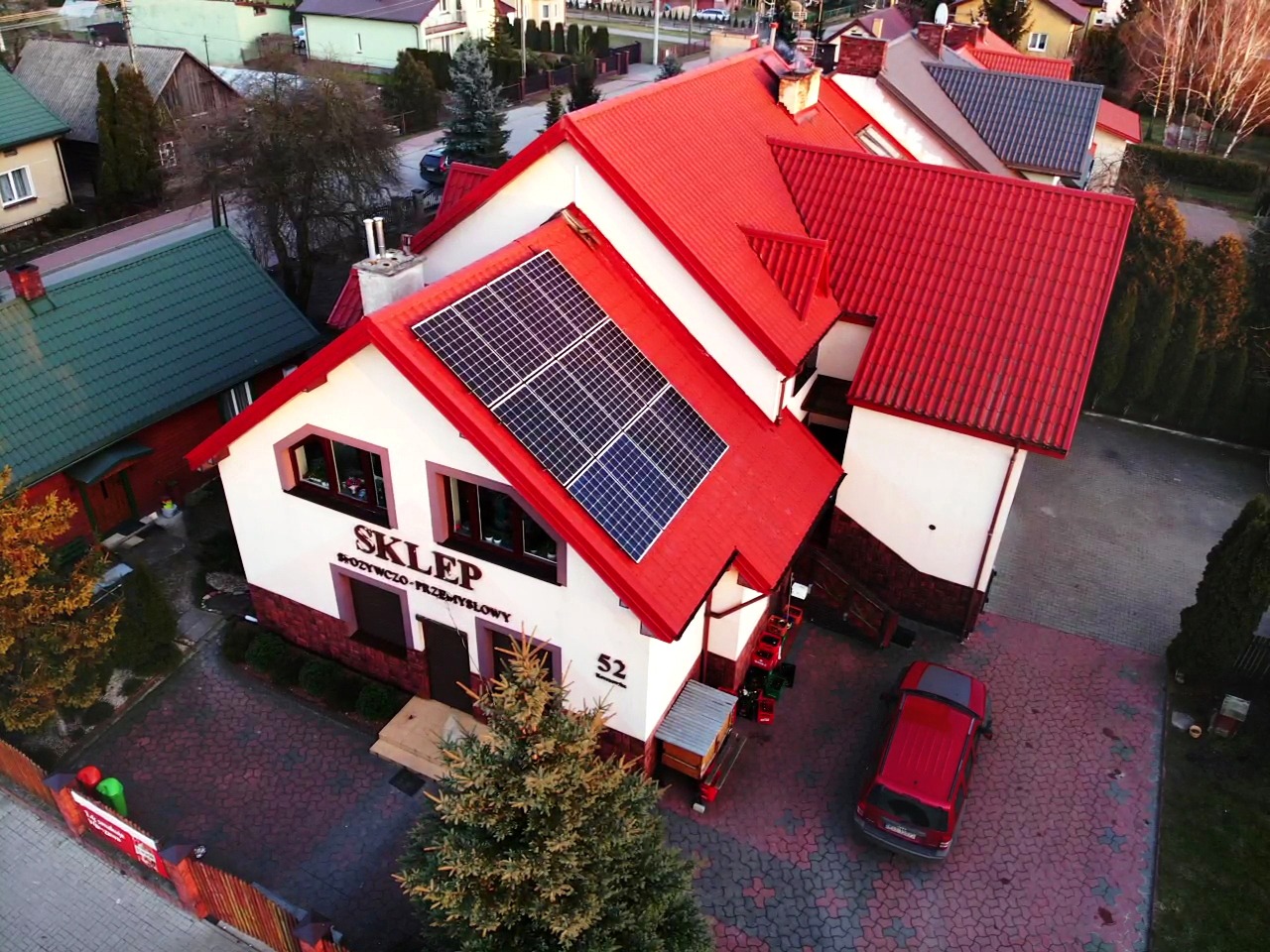 Widok z góry na budynek sklepu z czerwonym dachem i panelami słonecznymi na jednej z połaci, obok zaparkowany czerwony samochód.