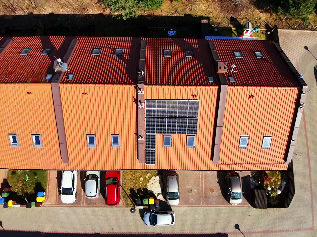Widok z góry na szereg domów z czerwonymi dachami, z panelami słonecznymi zamontowanymi na jednym z dachów, oraz zaparkowanymi samochodami przed budynkami.