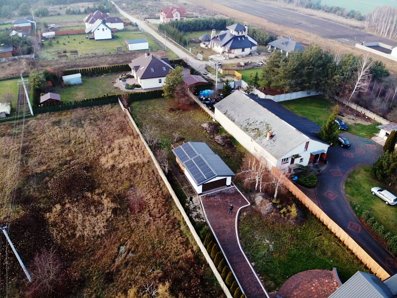 Widok z drona na dom z panelami słonecznymi na dachu garażu, otoczony ogrodem i innymi budynkami mieszkalnymi w słoneczny dzień.