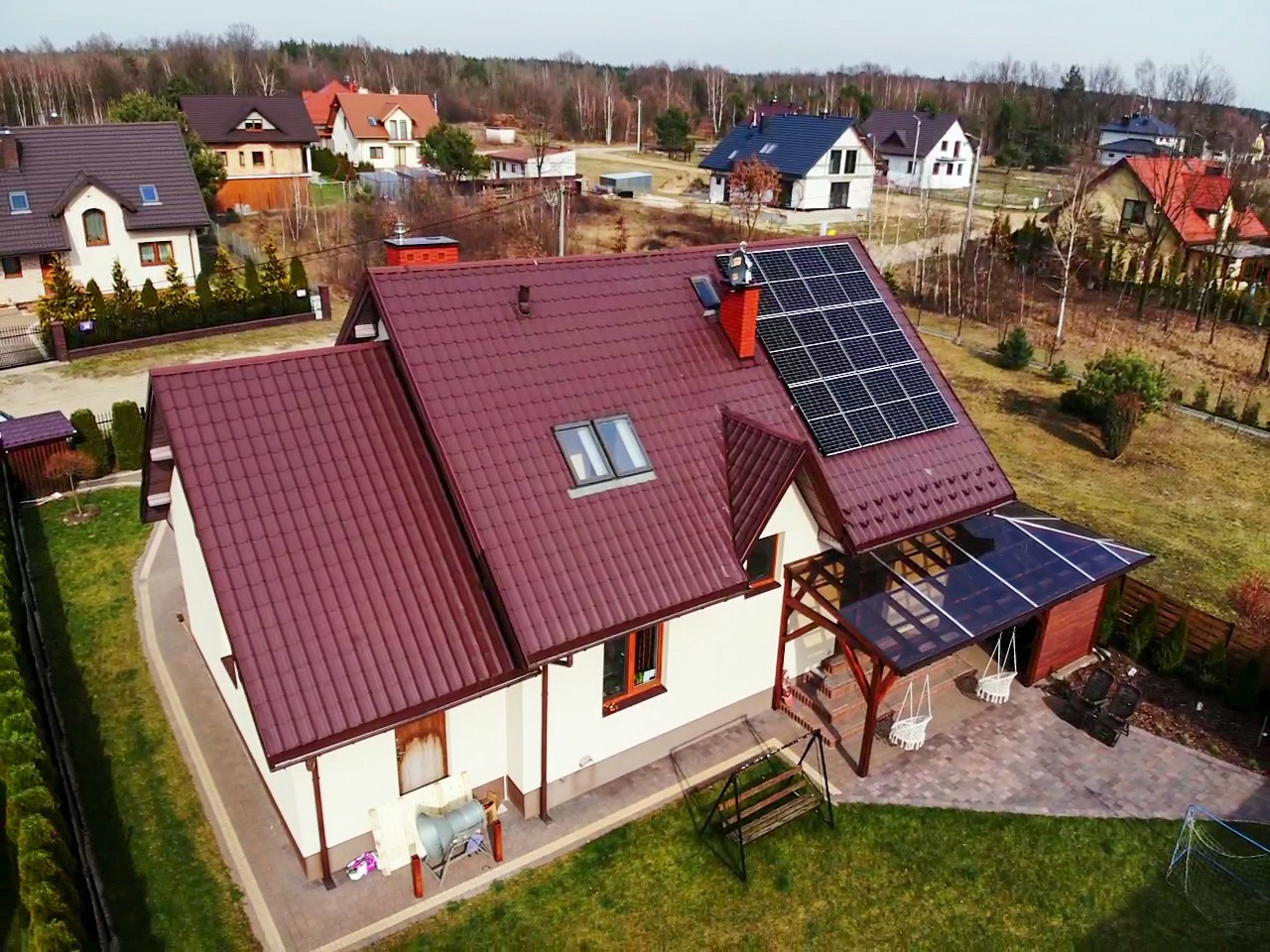 Widok z góry na dom jednorodzinny z panelami słonecznymi na dachu o ceglastym kolorze, przydomowa altana z huśtawkami i zadbany trawnik.