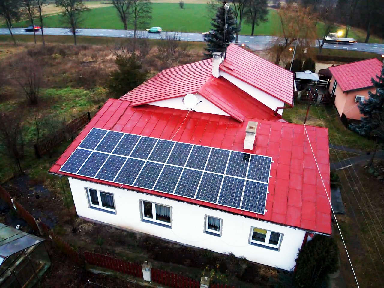 Widok z góry na dom z czerwoną dachówką i panelami słonecznymi na dachu, widoczna droga i otoczenie w jesiennej scenerii.