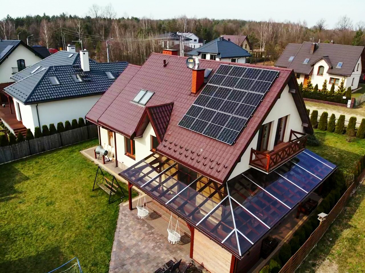 Dom jednorodzinny z brązowym dachem i panelami fotowoltaicznymi na połaci dachu, widok z lotu ptaka, oszklona weranda, zadbany trawnik.