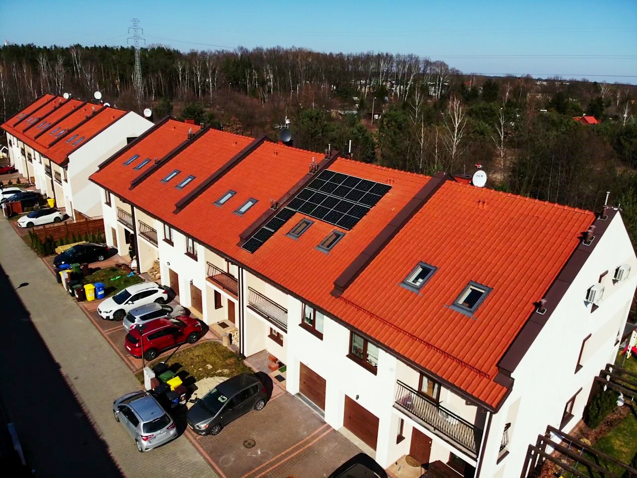 Widok z góry na szereg domów jednorodzinnych z czerwonymi dachami i panelami słonecznymi na niektórych dachach, zaparkowane samochody przed budynkami.