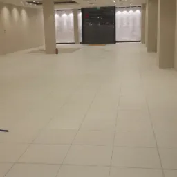 Przestronne, puste pomieszczenie biurowe z jasnymi, kwadratowymi płytkami podłogowymi, kolumnami nośnymi i zamkniętą, metalową bramą rolowaną w tle, oświetlone równomiernie przez wbudowane lampy...