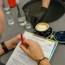 Sędzia ocenia cappuccino z wzorem serca podczas konkursu baristów, trzymając arkusz ocen i czerwony ołówek, obok szklanki wody, butelki wody i stopera.