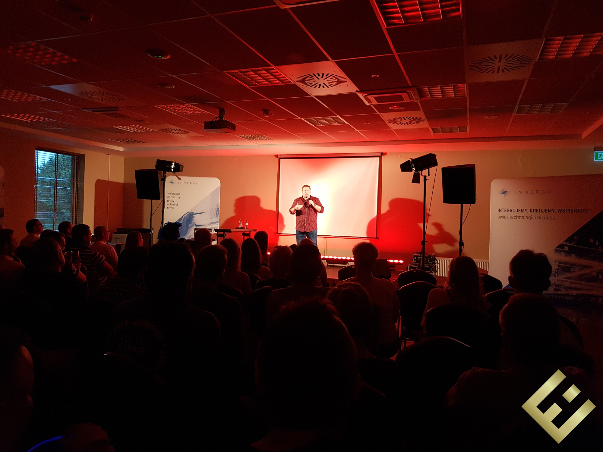 Prelegent w czerwonej koszuli przemawia do publiczności podczas konferencji w Krakowie, oświetlonej czerwonym światłem scenicznym, z widocznymi banerami firmowymi i rzędami krzeseł dla słuchaczy.