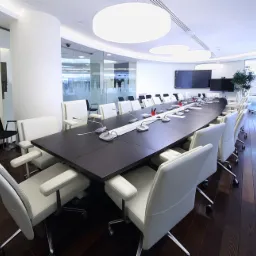 Nowoczesna sala konferencyjna z długim, ciemnym stołem, białymi skórzanymi krzesłami na kółkach, mikrofonami i oświetleniem LED, parkiet na podłodze.