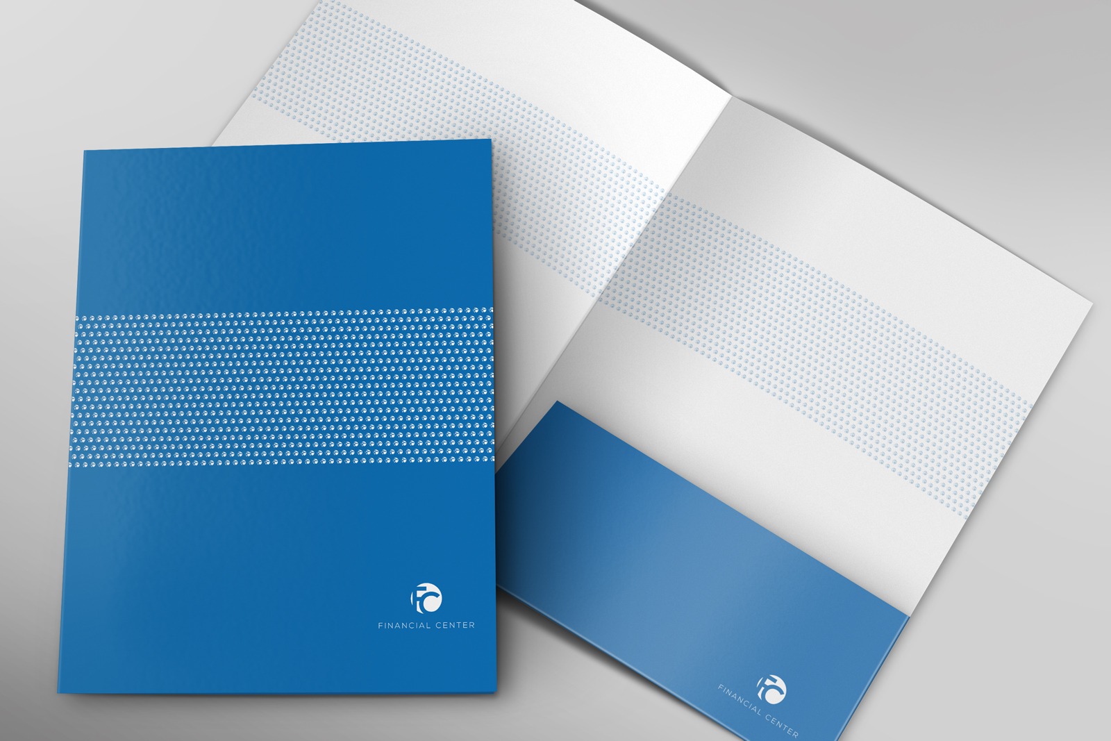 Niebieski folder firmowy Financial Center z białym logo i paskiem z drobnymi kropkami, otwarty obok zamkniętego, prezentacja identyfikacji wizualnej.