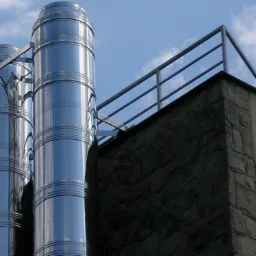 Dwa pionowe, segmentowe, metalowe kominy wentylacyjne na zewnątrz budynku z kamienną elewacją i metalową balustradą na dachu, widoczne na tle błękitnego nieba z chmurami.