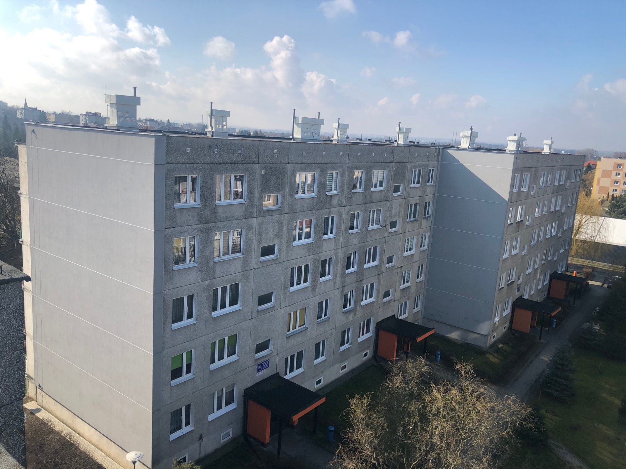Widok z góry na szary blok mieszkalny z widocznymi kominami wentylacyjnymi na dachu, osiedlowym terenem zielonym i fragmentem miasta w tle, uchwycony w słoneczny dzień.