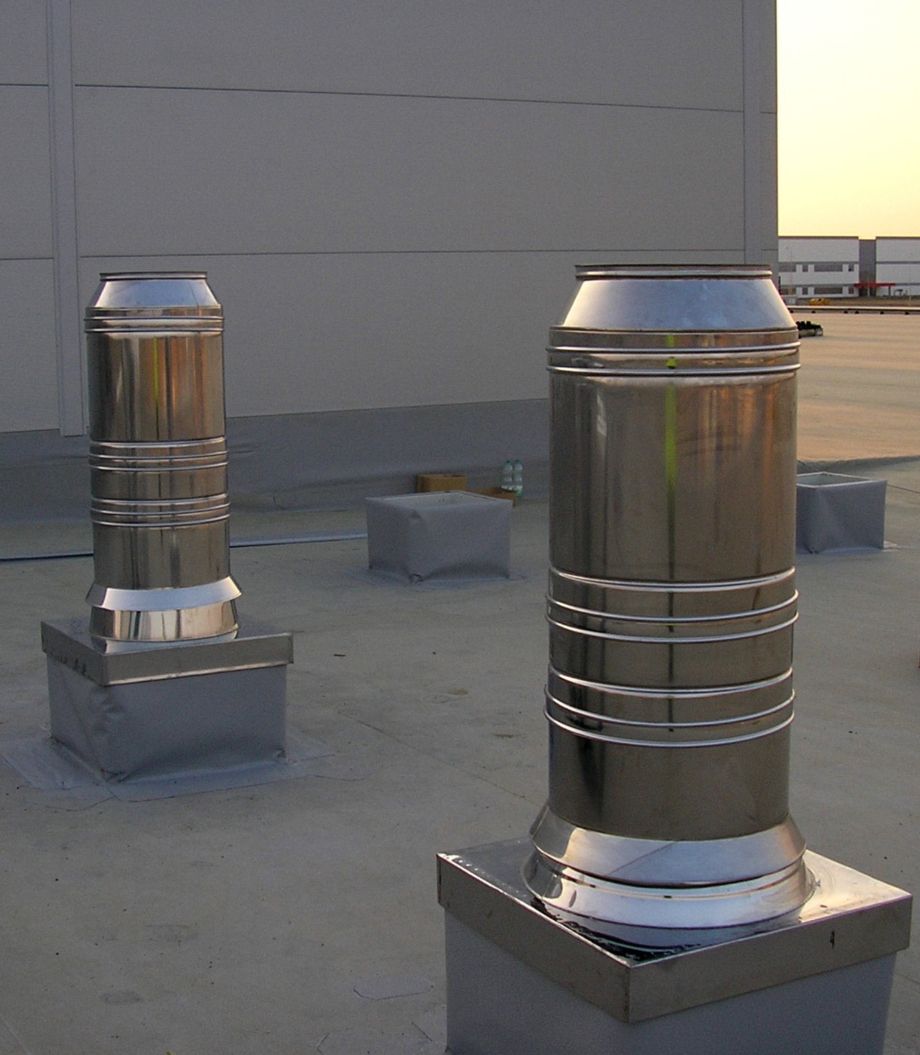 Dwa metalowe kominy wentylacyjne na płaskim dachu budynku przemysłowego, widoczne w oddali inne budynki, zachodzące słońce.