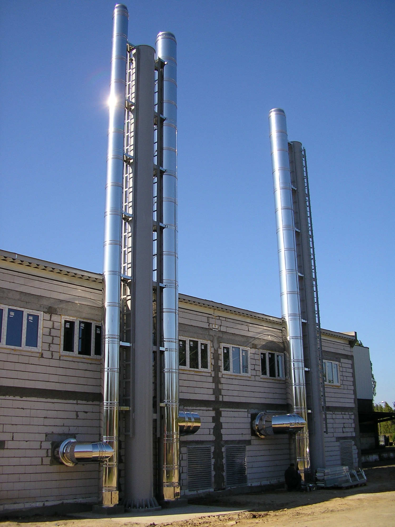 Błyszczące, metalowe kominy wentylacyjne zamontowane na zewnętrznej ścianie budynku z białej cegły, z widocznymi elementami mocującymi i drabinkami, na tle błękitnego nieba.