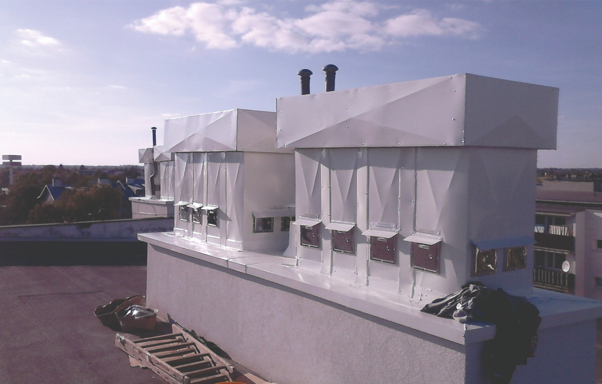Białe, metalowe obudowy systemów wentylacyjnych z otworami rewizyjnymi na dachu budynku, widoczne kominy wentylacyjne, drabina i ubrania robocze obok.