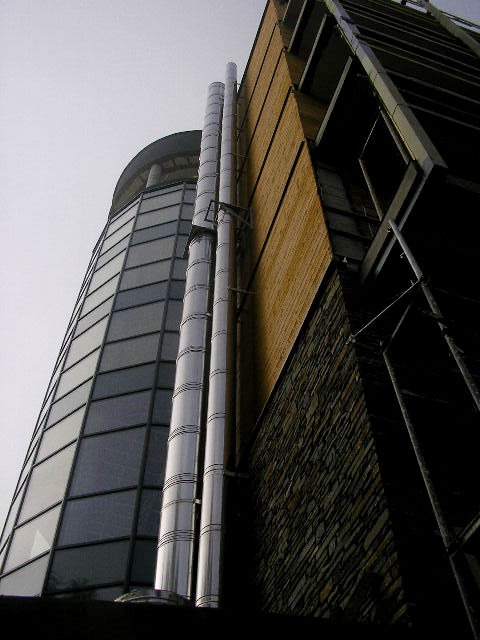 Dwa pionowe, metalowe przewody wentylacyjne na zewnątrz budynku biurowego z ceglaną ścianą i szklaną elewacją, widok z dołu.