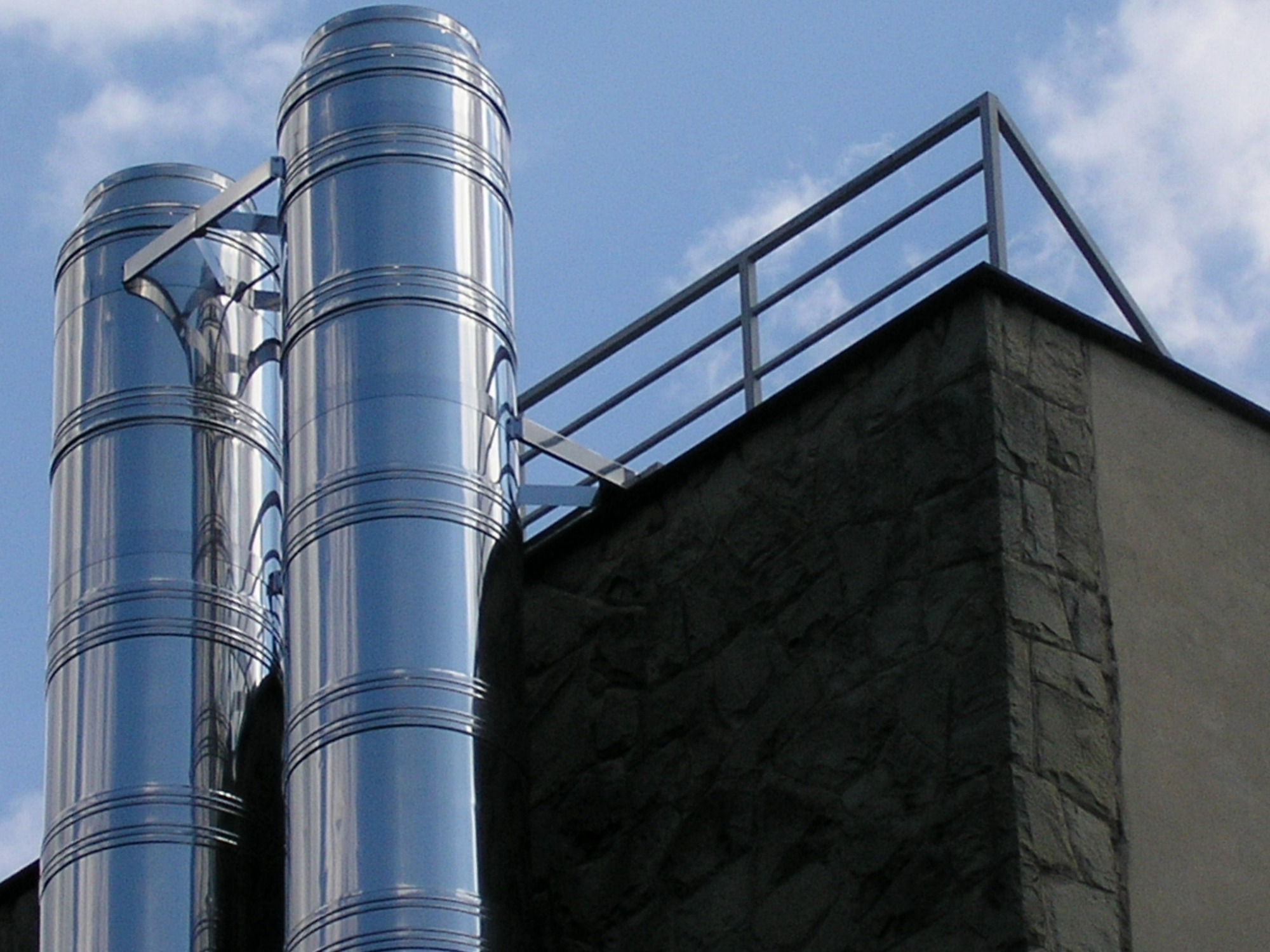 Dwa pionowe, segmentowe, metalowe kominy wentylacyjne na zewnątrz budynku z kamienną elewacją i metalową balustradą na dachu, widoczne na tle błękitnego nieba z chmurami.