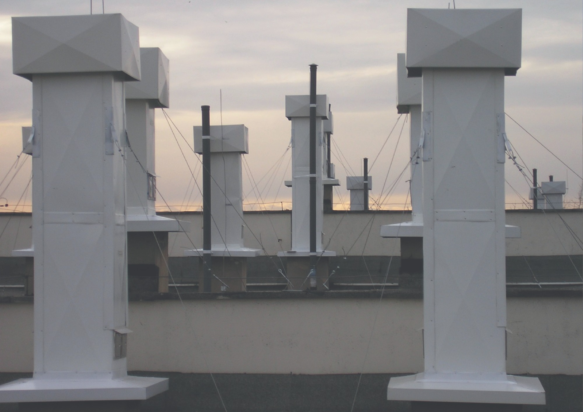 Widok na dachu z kilkoma białymi, kwadratowymi kominami wentylacyjnymi z czarnymi rurami wychodzącymi z niektórych z nich, zabezpieczonymi linkami, na tle pochmurnego nieba.