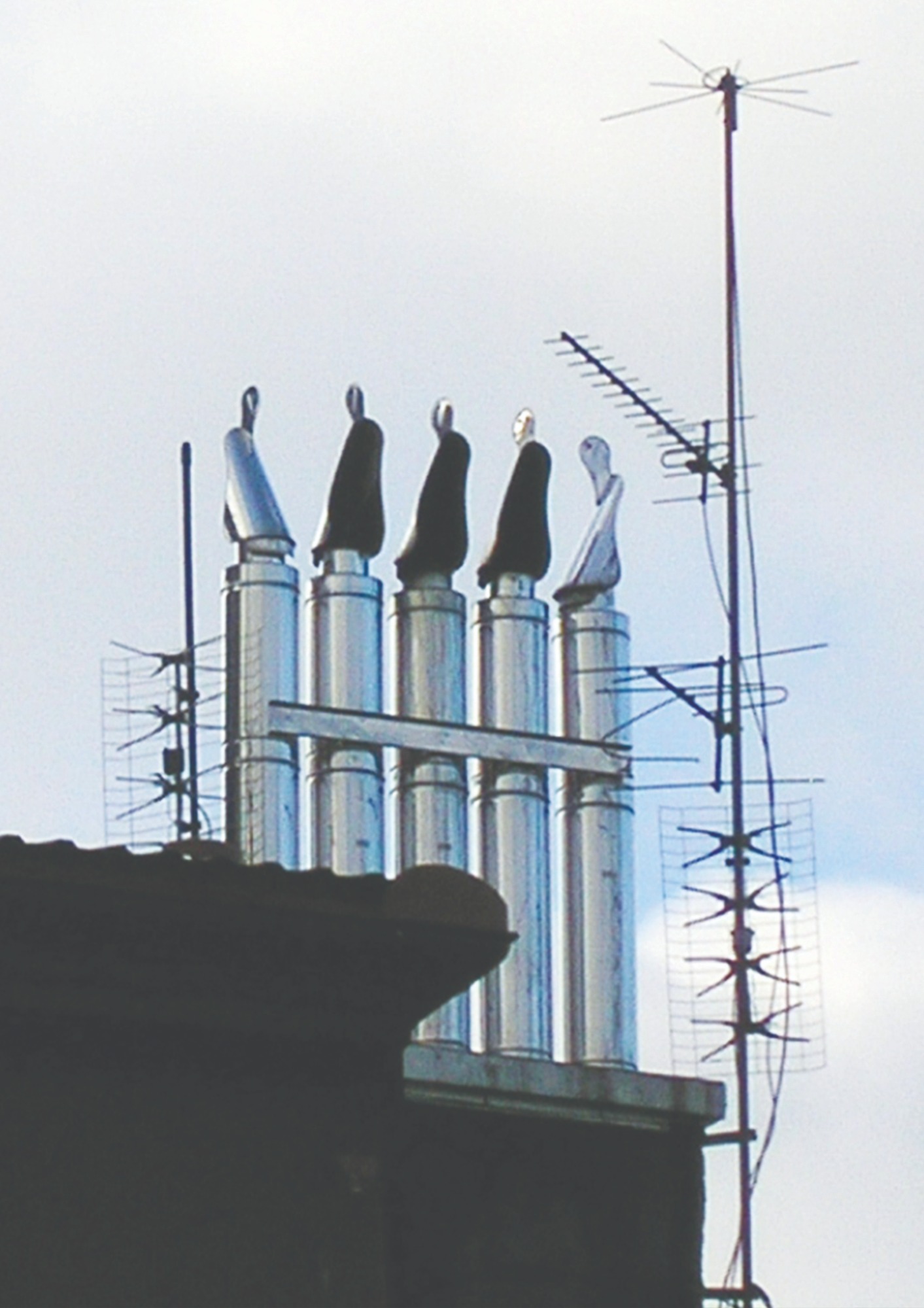 Szereg chromowanych kominów wentylacyjnych z daszkami na dachu budynku, obok anteny telewizyjnej.