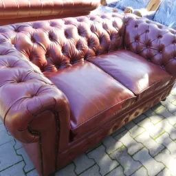 Dwuosobowa sofa w stylu Chesterfield, obita czerwoną skórą, z charakterystycznym pikowaniem, sfotografowana na zewnątrz na tle kostki brukowej z widocznymi chwastami.