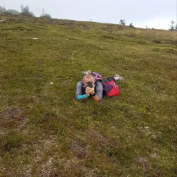 Mężczyzna leżący na trawiastym zboczu góry, robiący zdjęcie telefonem, z plecakiem obok, w tle zalesione wzgórze spowite mgłą.
