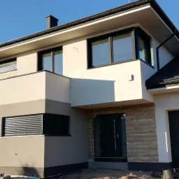 Nowoczesny dom jednorodzinny z elewacją w odcieniach beżu i szarości, czarne okna i dachówka, widoczne ślady po pracach wykończeniowych wokół budynku.