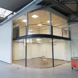 Nowoczesne biuro z przeszklonymi ścianami i antresolą, z widocznym krzesłem na środku i wózkiem transportowym z boku, oświetlone lampami sufitowymi.