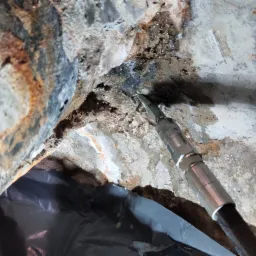 Wiertło wiertarki udarowej w otworze w wilgotnej, betonowej ścianie fundamentu z widocznymi śladami korozji i wykwitami solnymi.