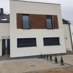 Nowoczesny dom jednorodzinny z jasną elewacją, drewnianymi akcentami okiennymi i szarą stolarką okienną, otoczony nowo wybrukowanym podjazdem i młodymi tujami.