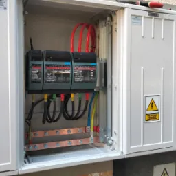 Otwarta szafka elektryczna z trzema zabezpieczeniami, przewodami w kolorach czerwonym, czarnym, zielonym i niebieskim, oraz miedzianą szyną uziemiającą. Na zamkniętych segmentach szafki naklejki...