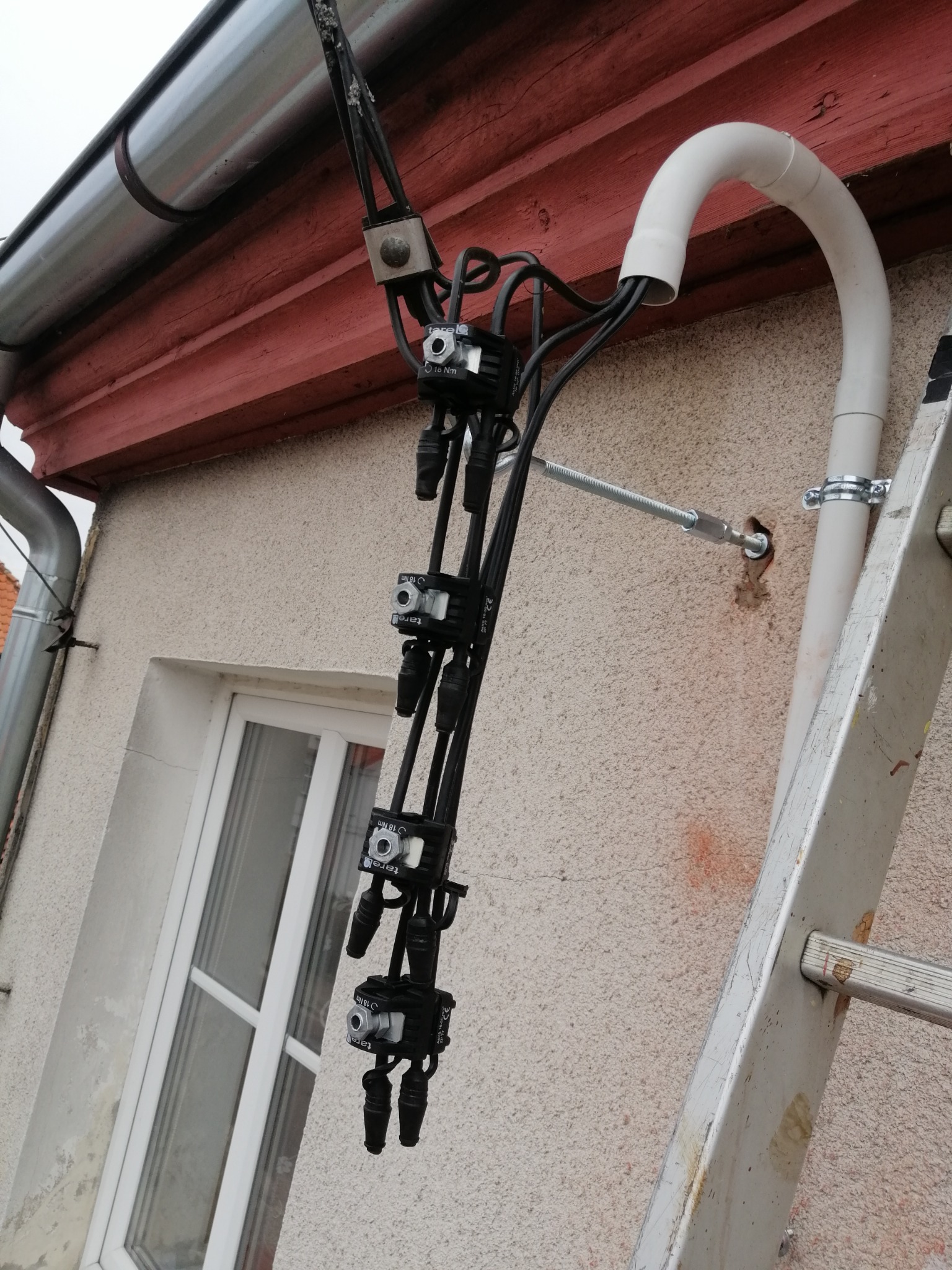 Zewnętrzna instalacja elektryczna z czarnymi kablami i złączkami, przymocowana do ściany budynku obok okna, zabezpieczona białym, giętkim peszlem, widoczna drabina oparta o ścianę.