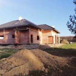 Surowy stan domu jednorodzinnego w trakcie budowy: widoczne ceglane ściany, dach z ciemnej dachówki, kominy i hałdy piasku na działce.