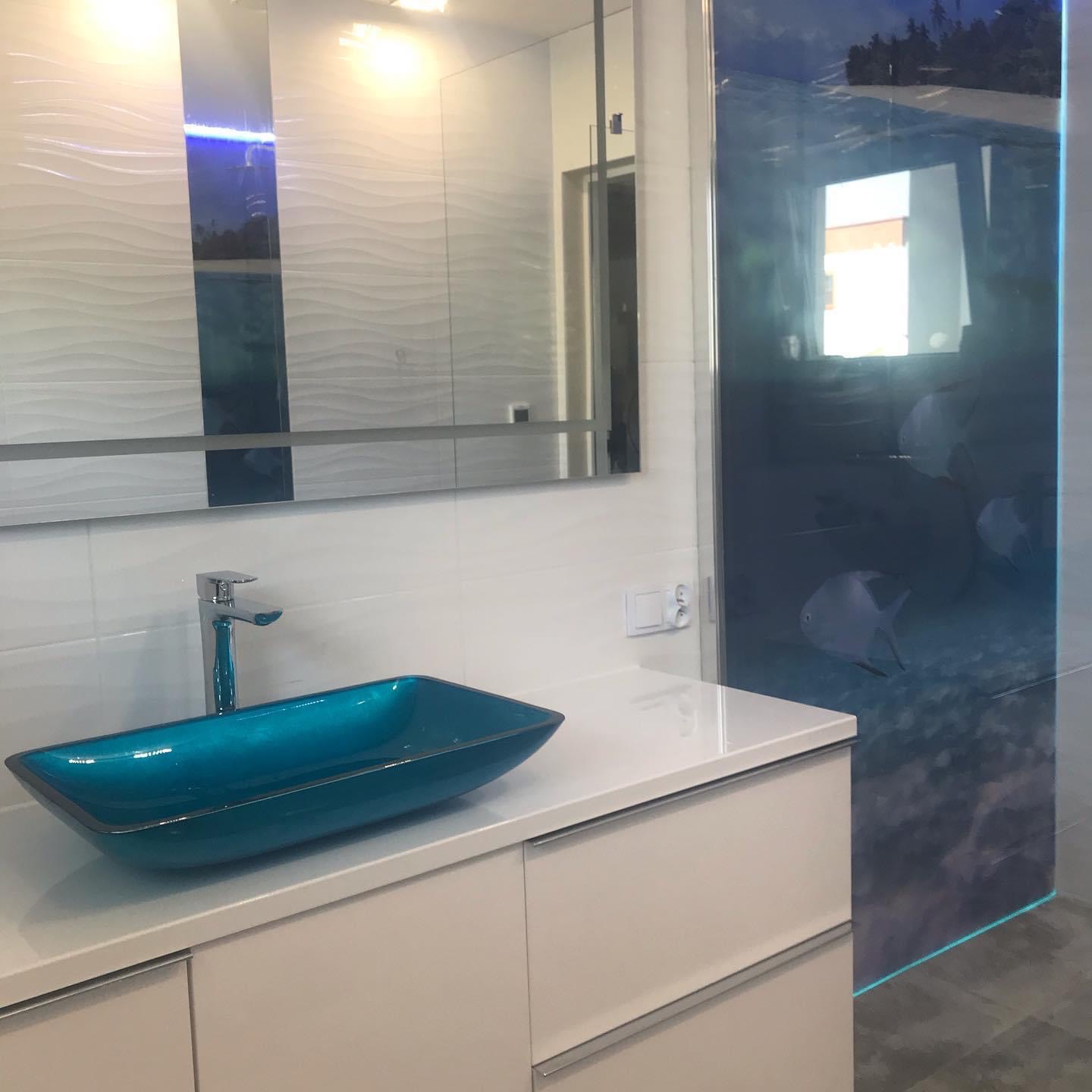 Nowoczesna łazienka z turkusową, szklaną umywalką nablatową, białą szafką pod umywalką, płytkami o falującej strukturze na ścianie oraz kabiną prysznicową z motywem podwodnego świata.