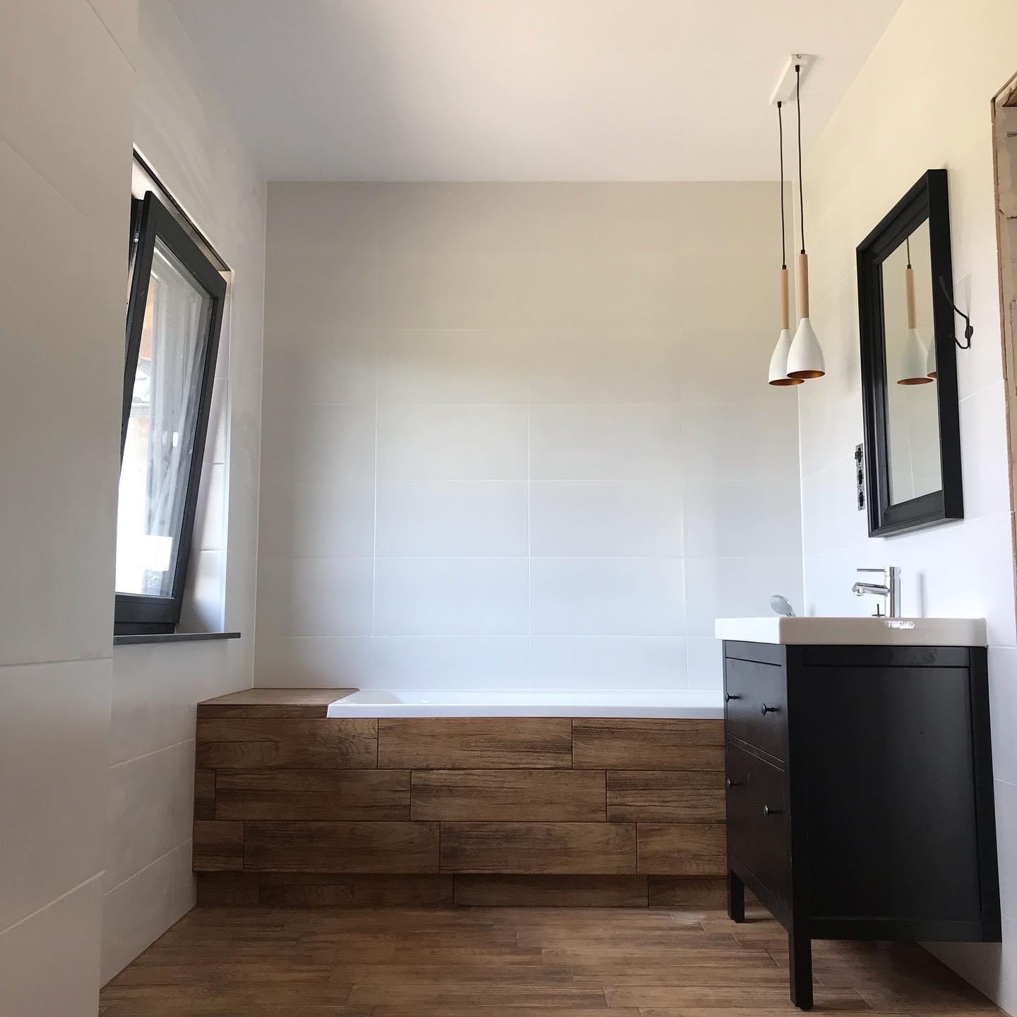 Nowoczesna łazienka z białą wanną obudowaną drewnem, czarna szafka pod umywalką, białe płytki na ścianach i podłoga imitująca drewno, okno w czarnej ramie.