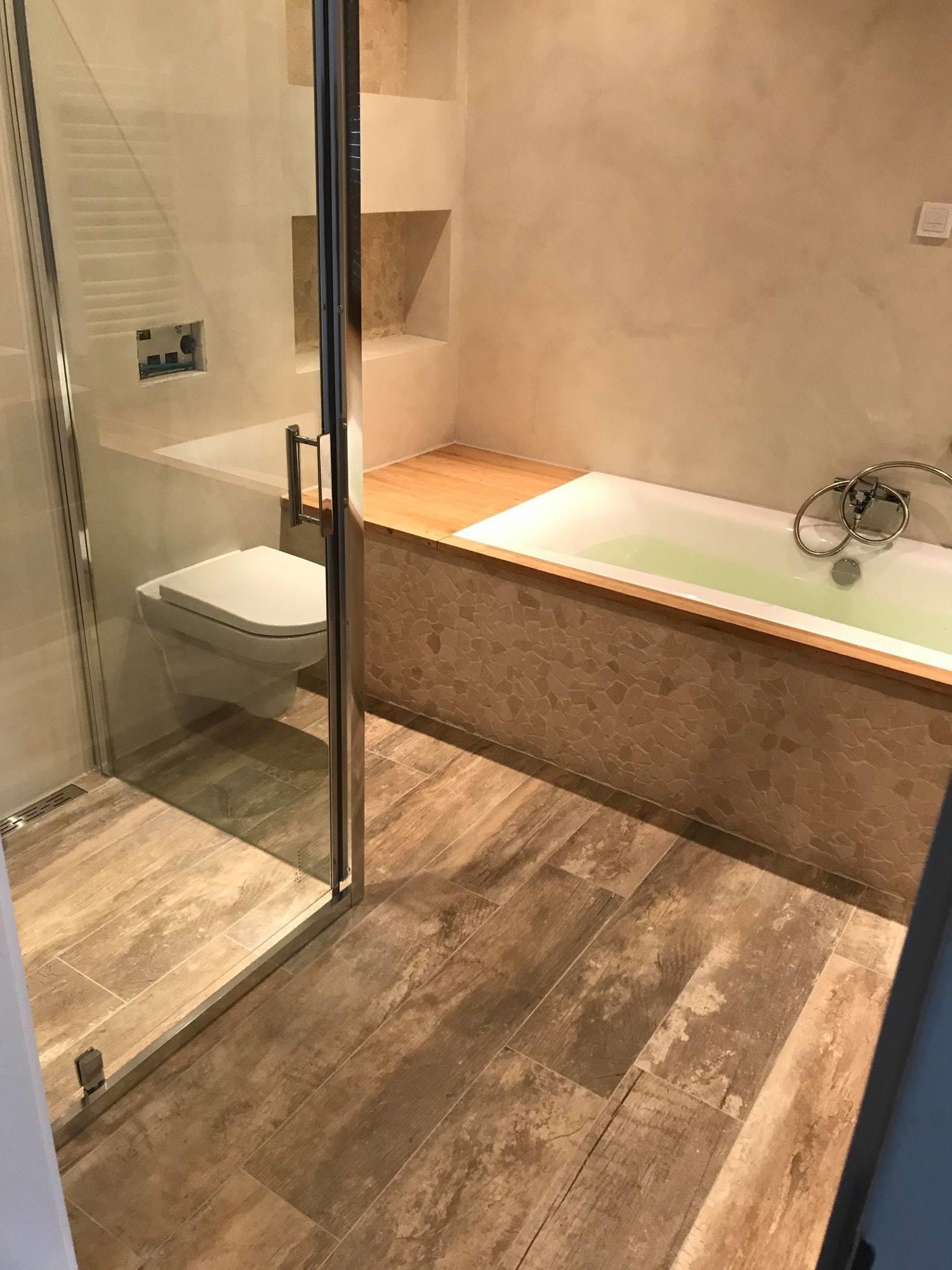 Wykończona łazienka z drewnopodobną podłogą, kabiną prysznicową ze szklanymi drzwiami, podwieszaną toaletą i obudowaną wanną z mozaiką oraz drewnianym blatem.