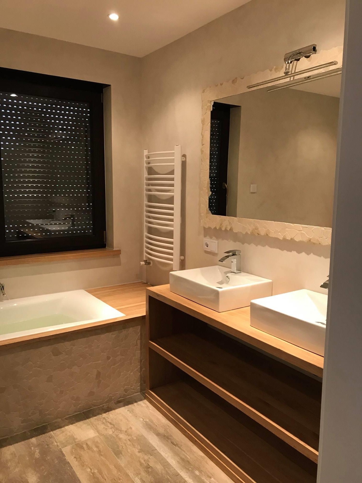 Nowoczesna łazienka z dwoma kwadratowymi umywalkami nablatowymi, drewnianym blatem, lustrem w ozdobnej ramie i grzejnikiem drabinkowym, z roletami w oknie.