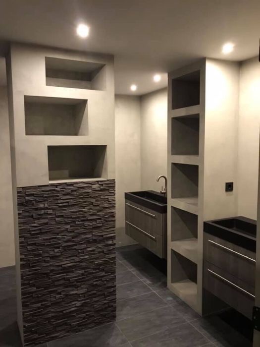 Nowoczesna łazienka z szarą, teksturowaną ścianą z wnękami, fragmentem ściany wyłożonym ciemnym kamieniem łupkowym, wiszącymi szafkami z umywalkami i półkami w zabudowie, oświetlona punktowymi...