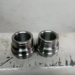 Dwa identyczne, metalowe elementy z widocznymi śladami obróbki skrawaniem, leżące na zabrudzonej, białej powierzchni.