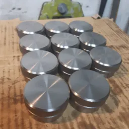 Partia okrągłych, metalowych elementów z radełkowanym brzegiem, ułożonych na drewnianej powierzchni, z fragmentem zielonej maszyny w tle.