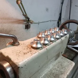 Rząd srebrnych, obrobionych na tokarce elementów na blacie tokarki z widocznymi śladami użytkowania i zieloną lampką w tle.