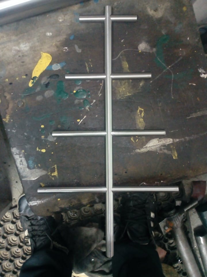 Metalowa konstrukcja spawana z rurek, ułożona na brudnym stole warsztatowym, widoczne buty robocze w dolnej części kadru.