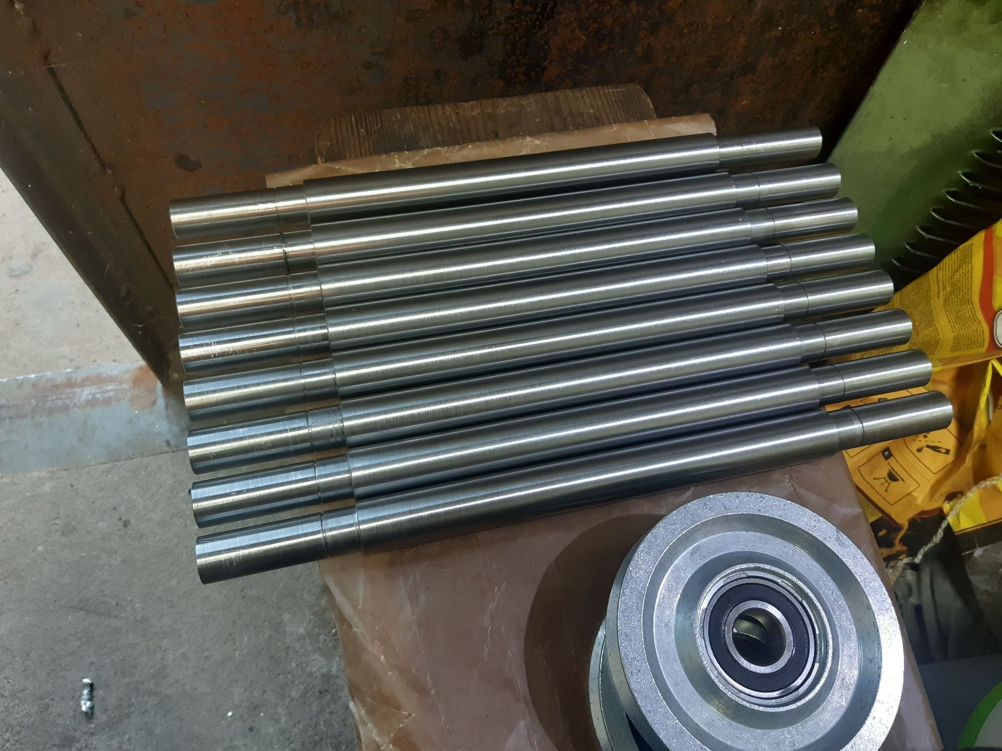 Stos równo ułożonych, obrobionych, metalowych prętów cylindrycznych o gładkiej powierzchni, leżących obok metalowego koła pasowego z łożyskiem kulkowym.