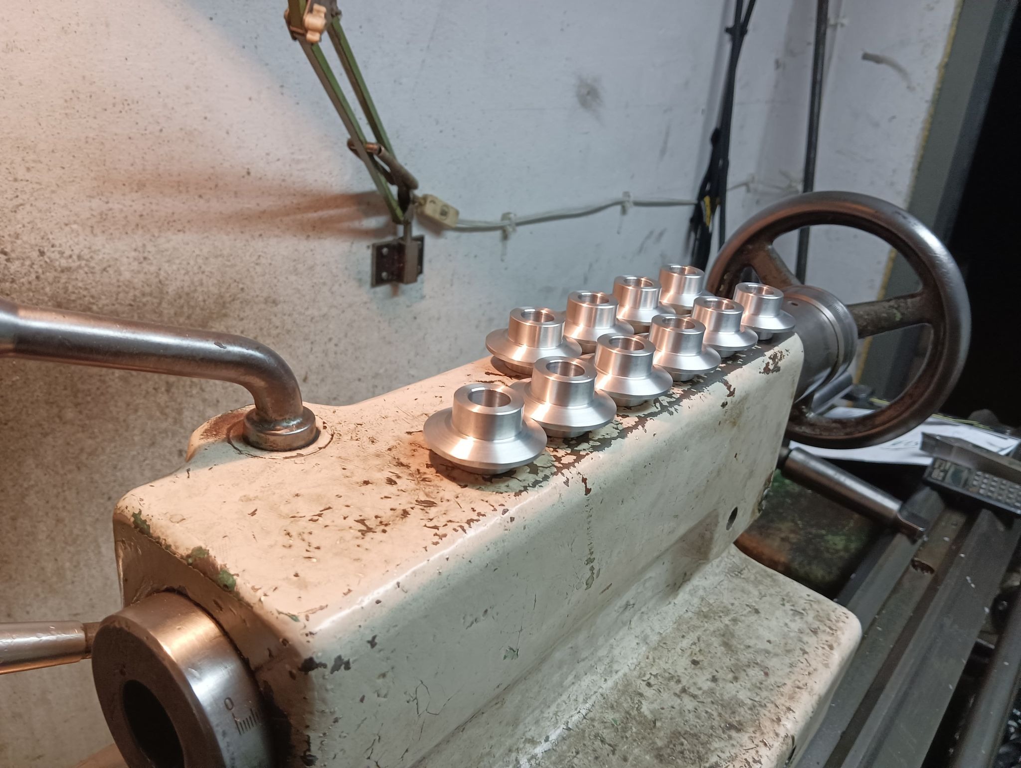 Rząd srebrnych, obrobionych na tokarce elementów na blacie tokarki z widocznymi śladami użytkowania i zieloną lampką w tle.