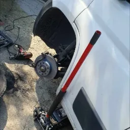 Biały samochód dostawczy na podnośniku hydraulicznym z odsłoniętą tarczą hamulcową, obok narzędzia warsztatowe i brudna szmata.