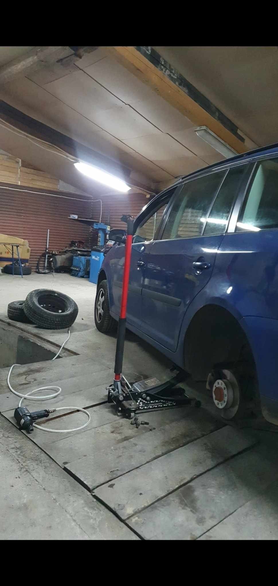 Niebieskie kombi podniesione na podnośniku hydraulicznym w warsztacie, brak koła, obok leżą opony i klucz pneumatyczny z przewodem.