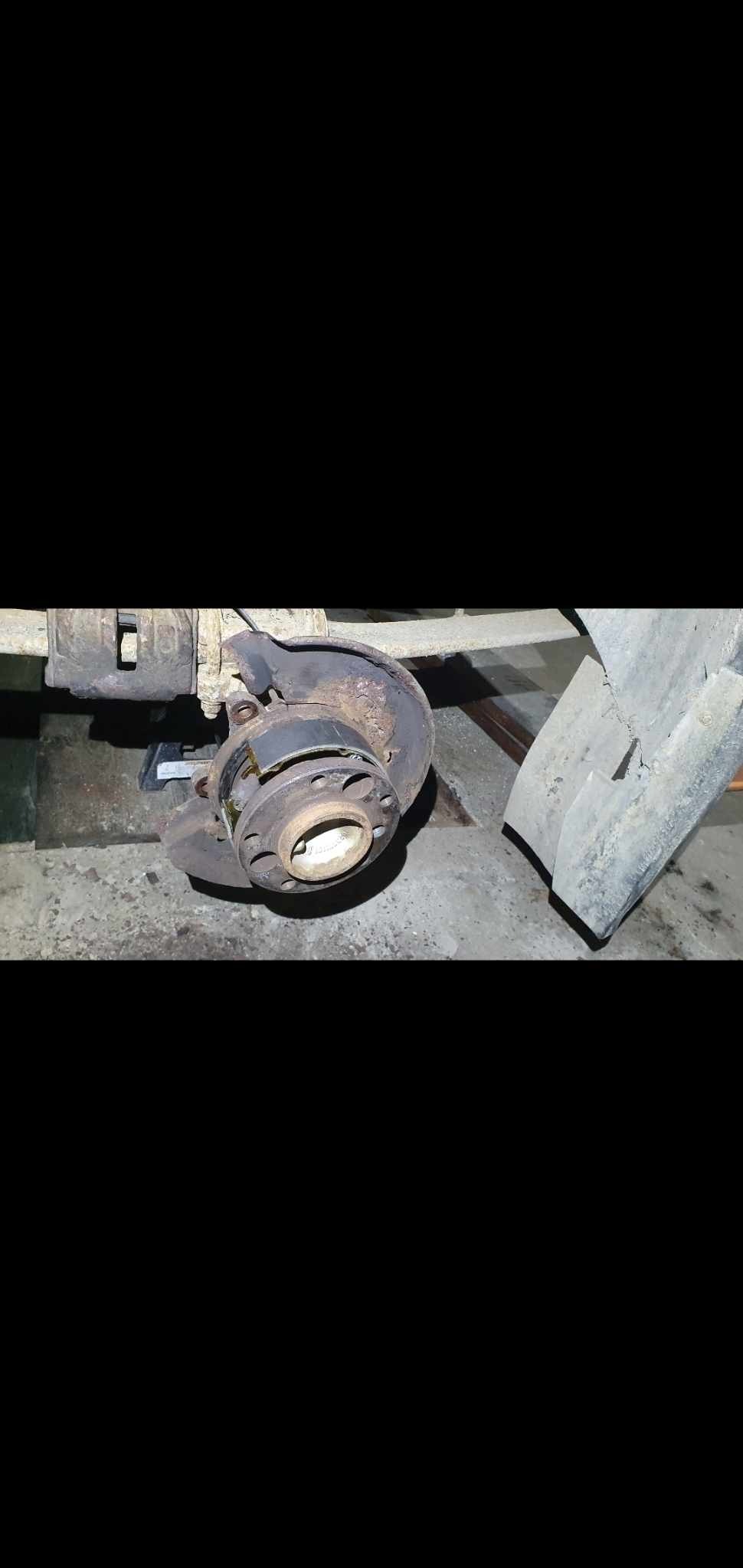 Zdjęcie przedstawia zdemontowane, skorodowane łożysko koła samochodowego z elementami zawieszenia, umieszczone na podnośniku w warsztacie, przy słabym oświetleniu.